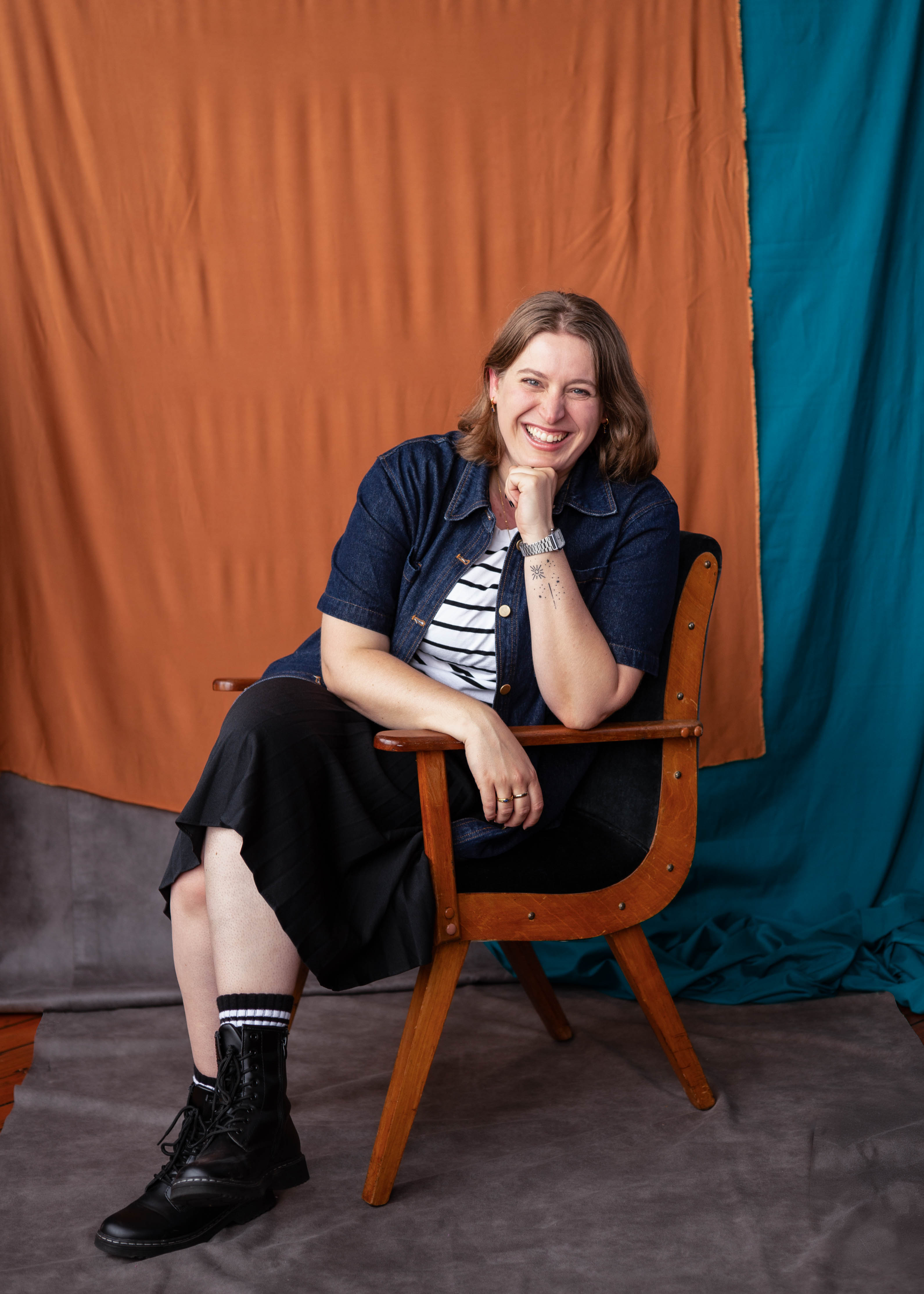 A picture of Jessica sitting in a wooden armchair against an orange and blue fabric background. They are looking towards the camera and laughing.